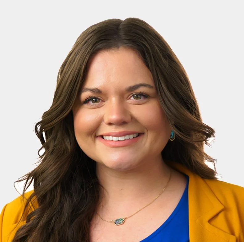 A woman with long, wavy brown hair and light skin smiling, wearing a yellow blazer, a blue top, teal earrings, and a gold necklace with a blue gemstone.