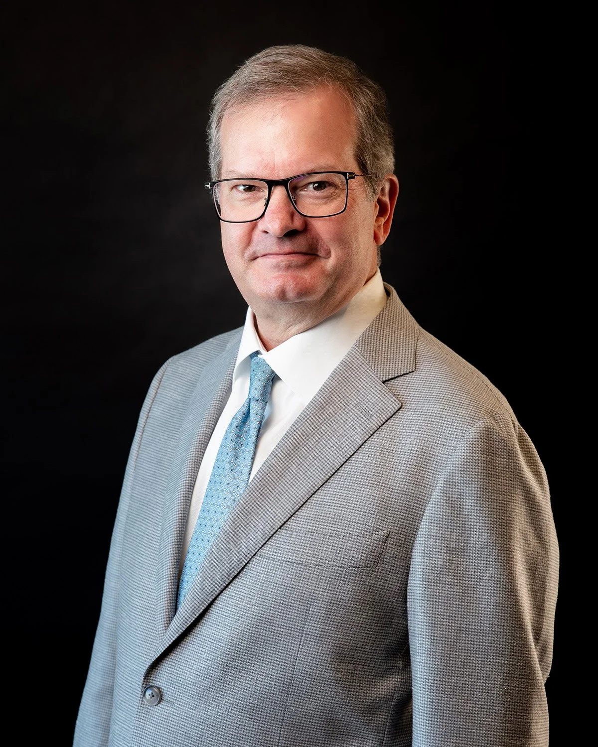 Headshot of a middle-aged man in a black pinstripe suit, white shirt, and light blue tie, against a dark background.