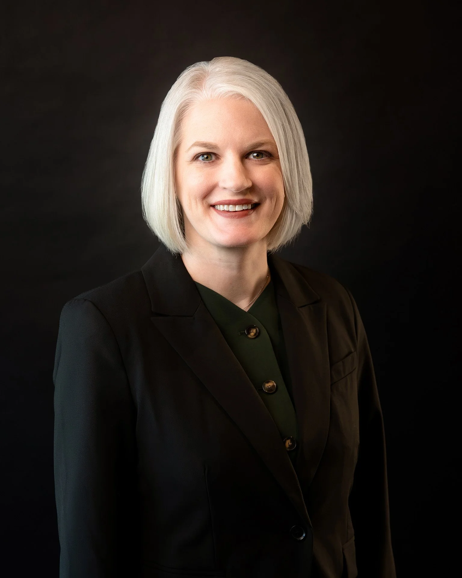 Headshot of a woman with short blonde hair, wearing a brown blazer and layered necklaces, smiling against a white background.