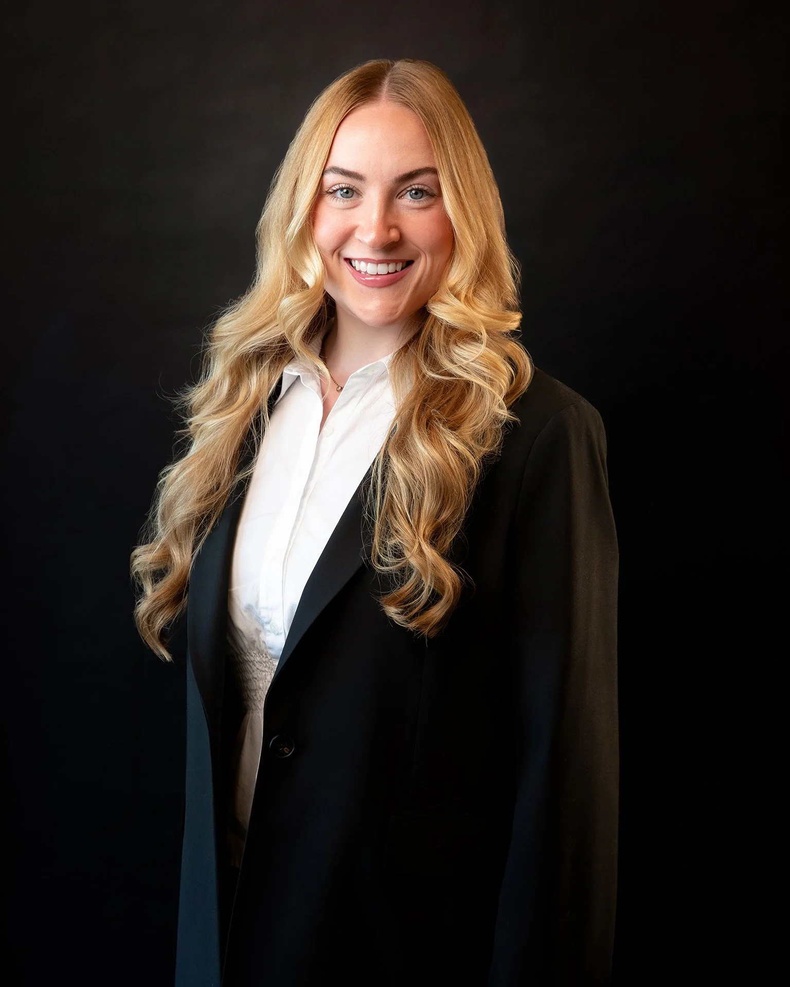 A young woman with long blonde hair smiling at the camera, wearing a teal satin shirt, against a plain light background.