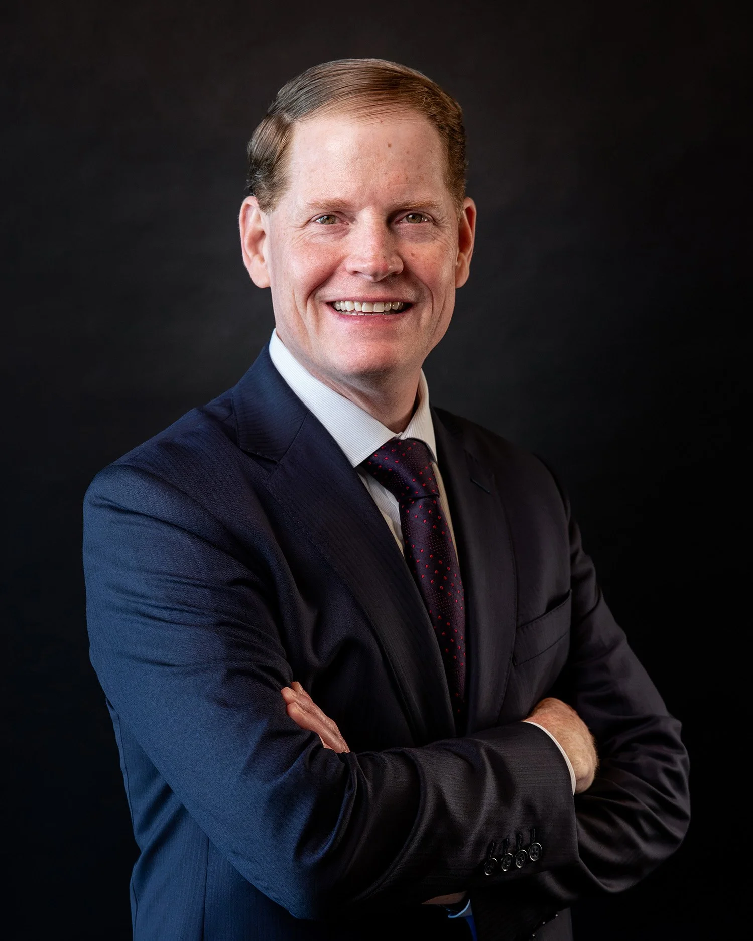 A smiling man in a dark business suit, light blue dress shirt, and patterned tie, with short reddish hair against a plain light background.