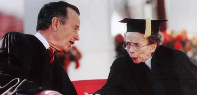 A man and an elderly woman in graduation attire exchanging a handshake or conversation outdoors.