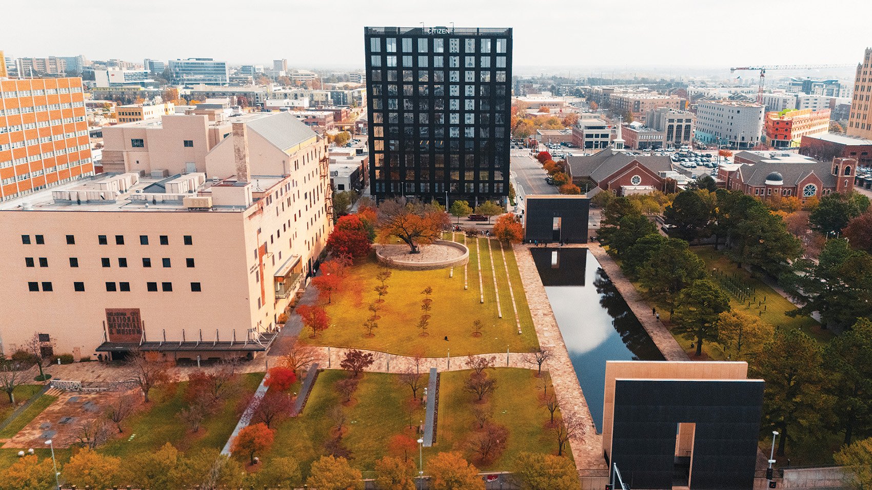 Aerial view of a city park with trees showing fall foliage, a water feature, and modern buildings surrounding it, including a tall black office building, a church with a steeple, and various commercial structures.
