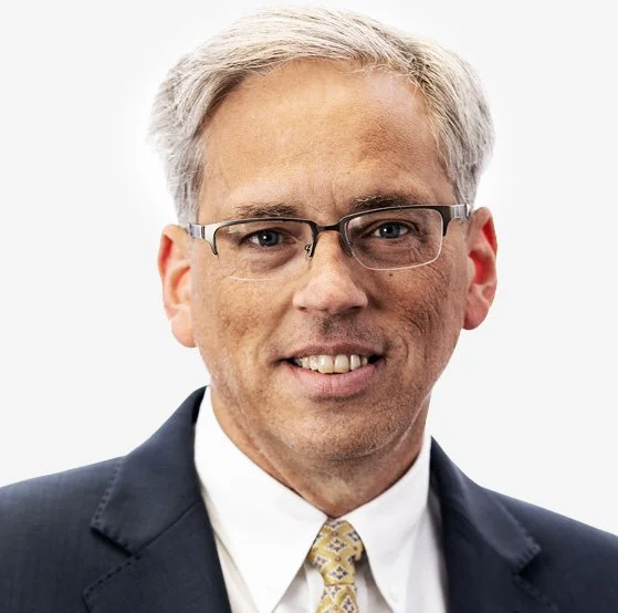 A smiling middle-aged man with gray hair, glasses, wearing a dark suit, white shirt, and patterned tie, portrait against a plain white background.