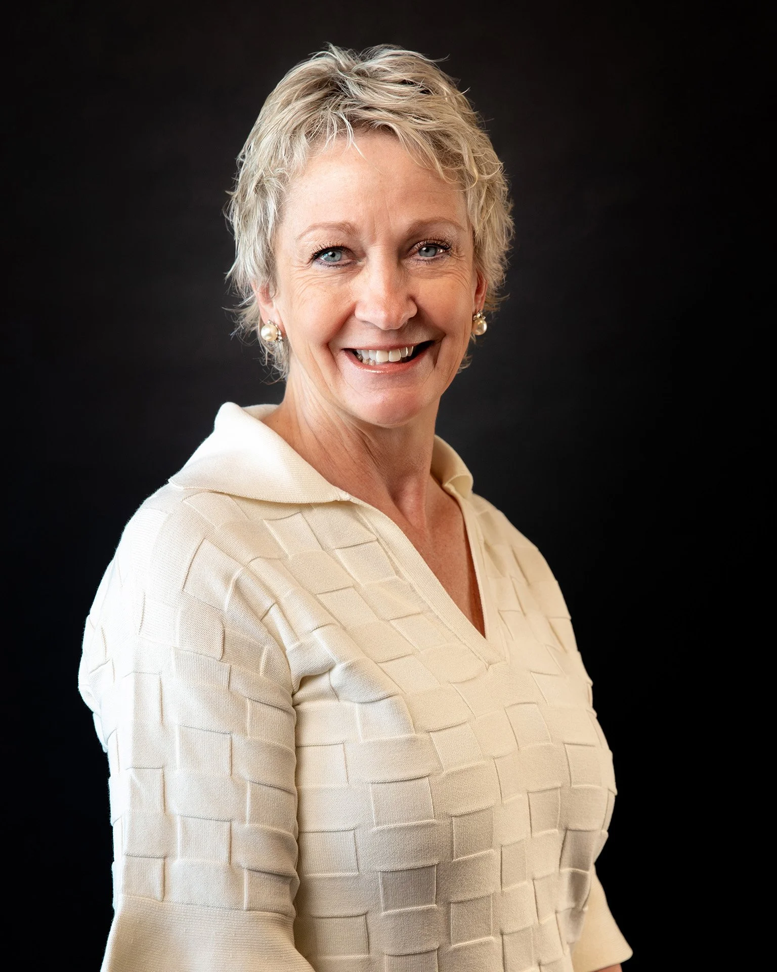 A professional woman with short blonde hair, wearing a black blazer over a white blouse, and gold hoop earrings, against a dark background.