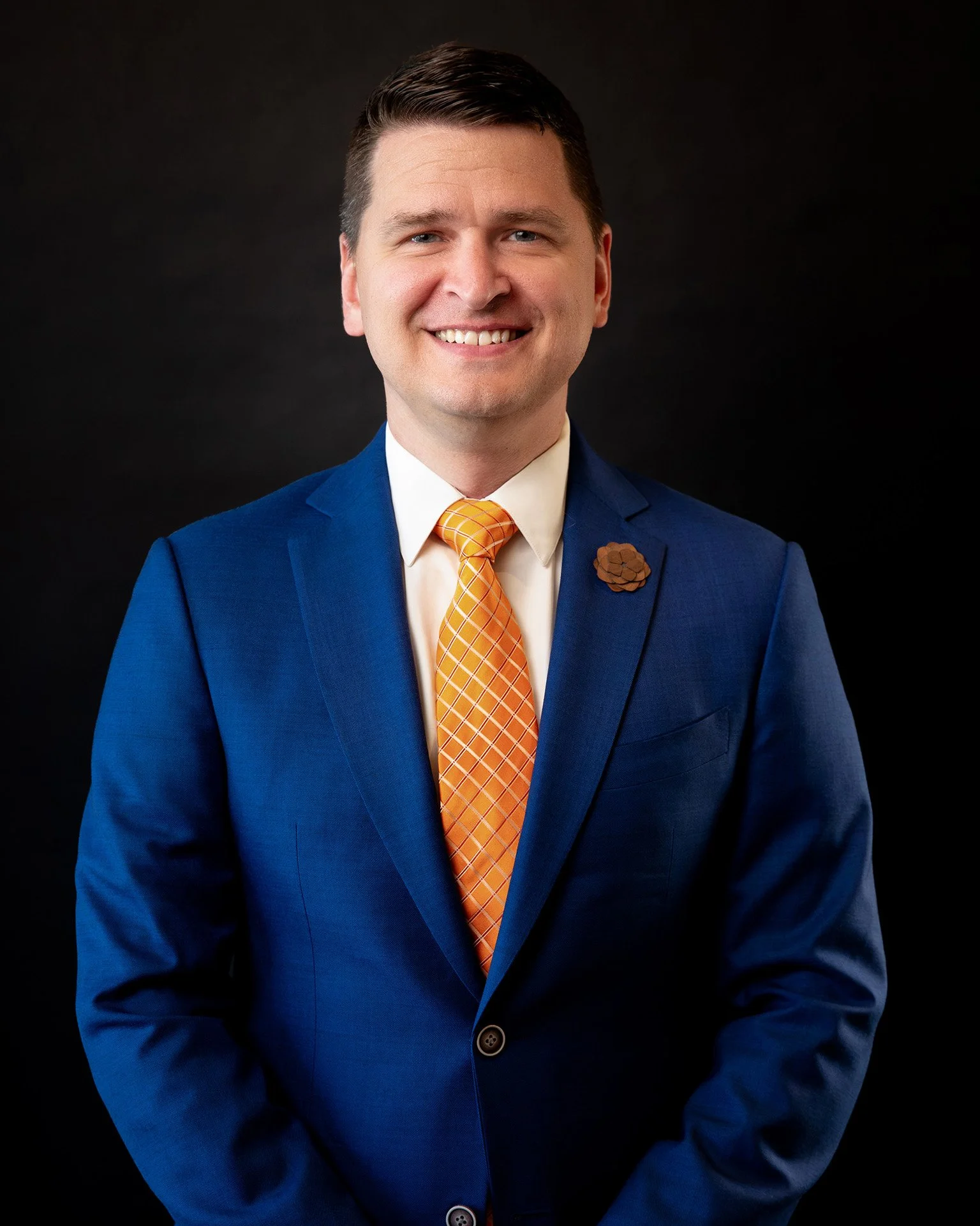 A smiling man in a suit with a white shirt and a yellow and blue striped tie, wearing a lapel pin. He has short dark hair and is standing against a plain white background.