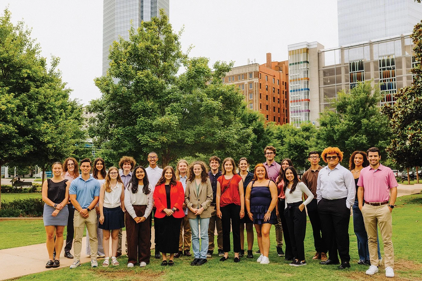 A diverse group of young adults standing outdoors in a city park surrounded by trees, with tall buildings in the background.