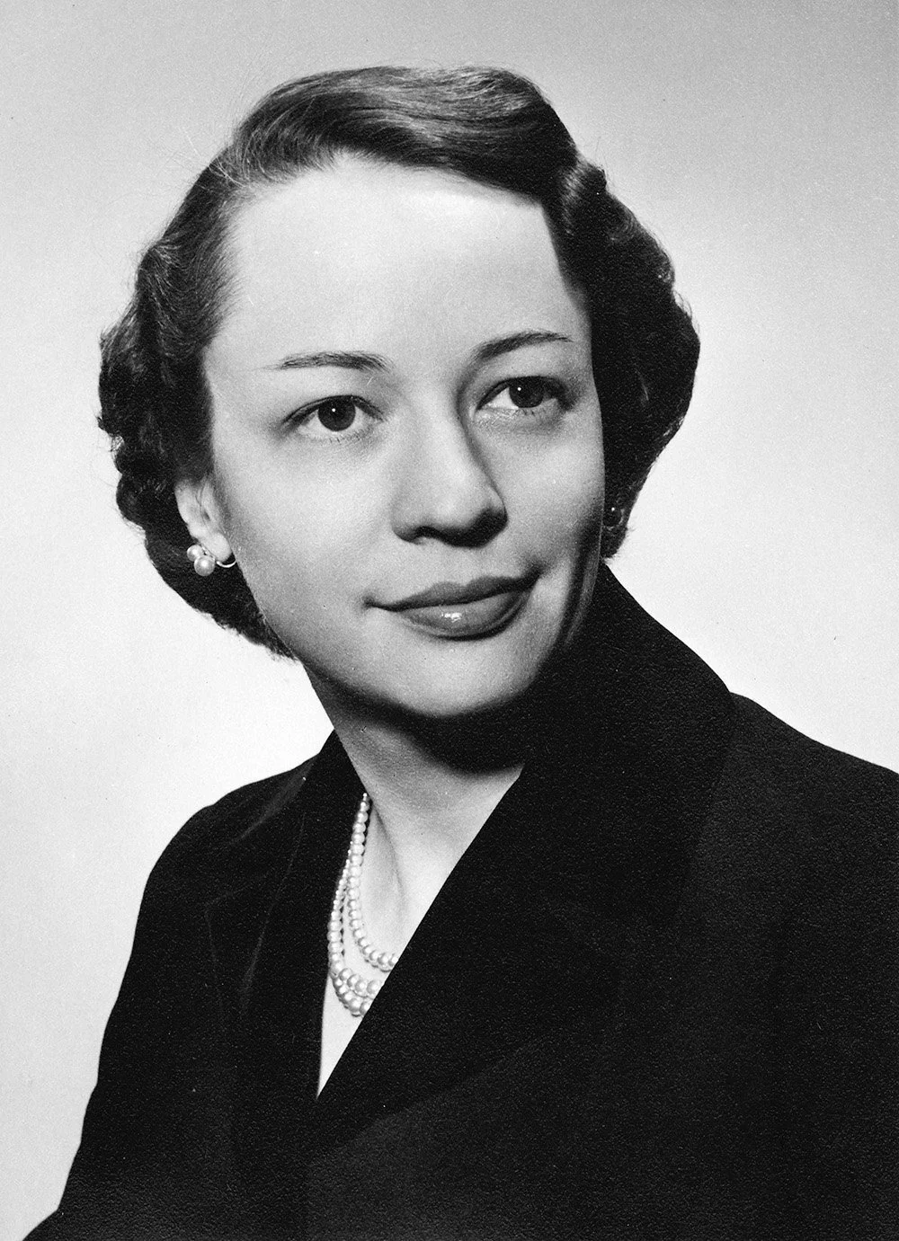 Black and white portrait of a woman with short dark wavy hair, wearing pearl earrings and a pearl necklace, dressed in a dark blazer, looking slightly to the side with a slight smile.