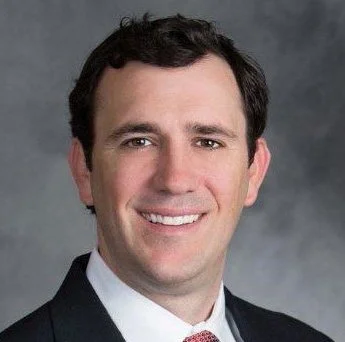 A professional headshot of a man in a suit smiling against a gray background.