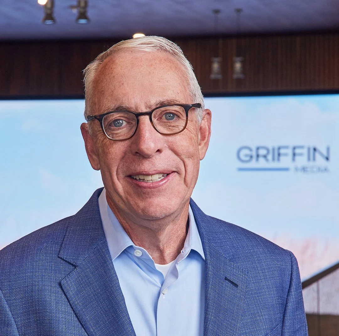 A smiling middle-aged man with short gray hair, glasses, wearing a blue suit jacket and light blue dress shirt, standing indoors with a projector screen in the background displaying the text 'GRIFFIN' and 'MEDIA'.