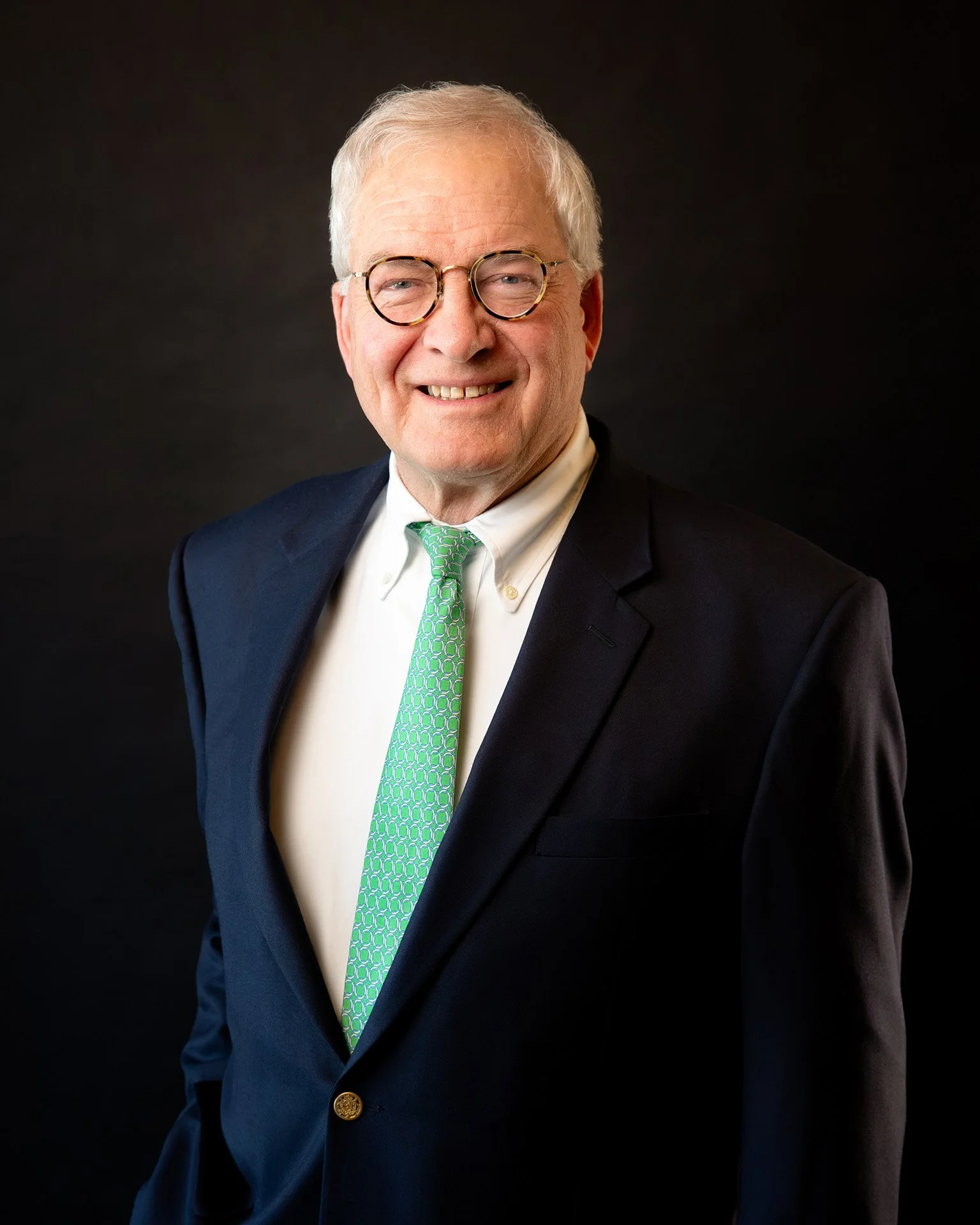 A smiling older man with glasses, gray hair, wearing a navy suit and a striped tie, against a white background.