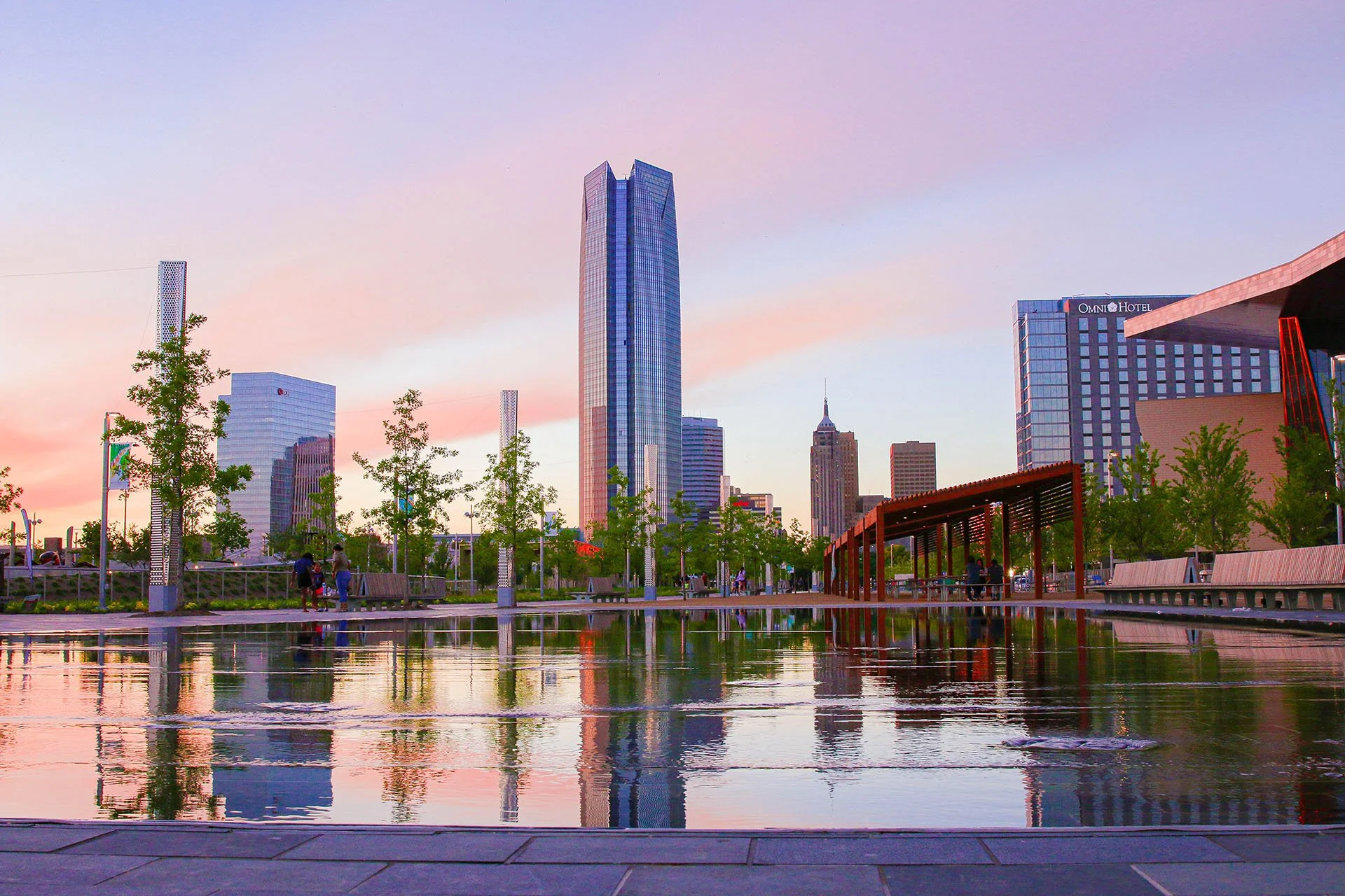 City skyline at sunset with tall skyscrapers, trees, a reflecting pool, and benches in a park.