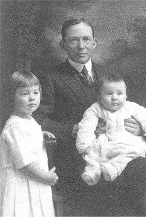 A man dressed in a suit sitting with two young girls, one standing and one sitting on his lap, in a black-and-white portrait.