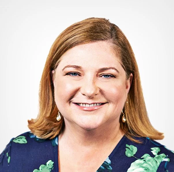 Portrait of a smiling woman with shoulder-length reddish-brown hair, wearing a dark blue blouse with green floral print and gold earrings, against a white background.