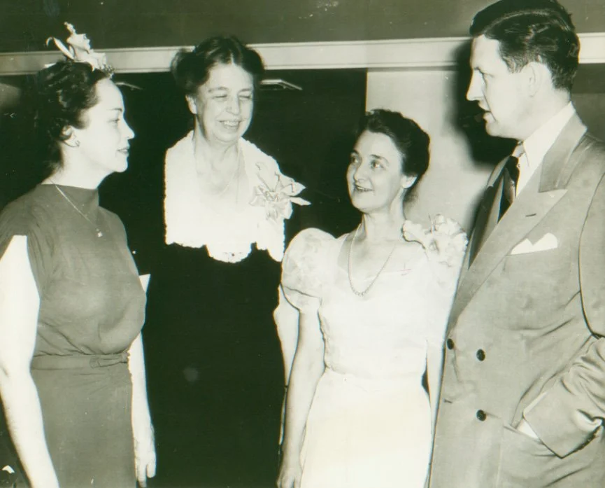 Four people, two women and one man, engaged in conversation in what appears to be a formal or semi-formal setting, possibly a social or professional event, with an older woman smiling in the center.