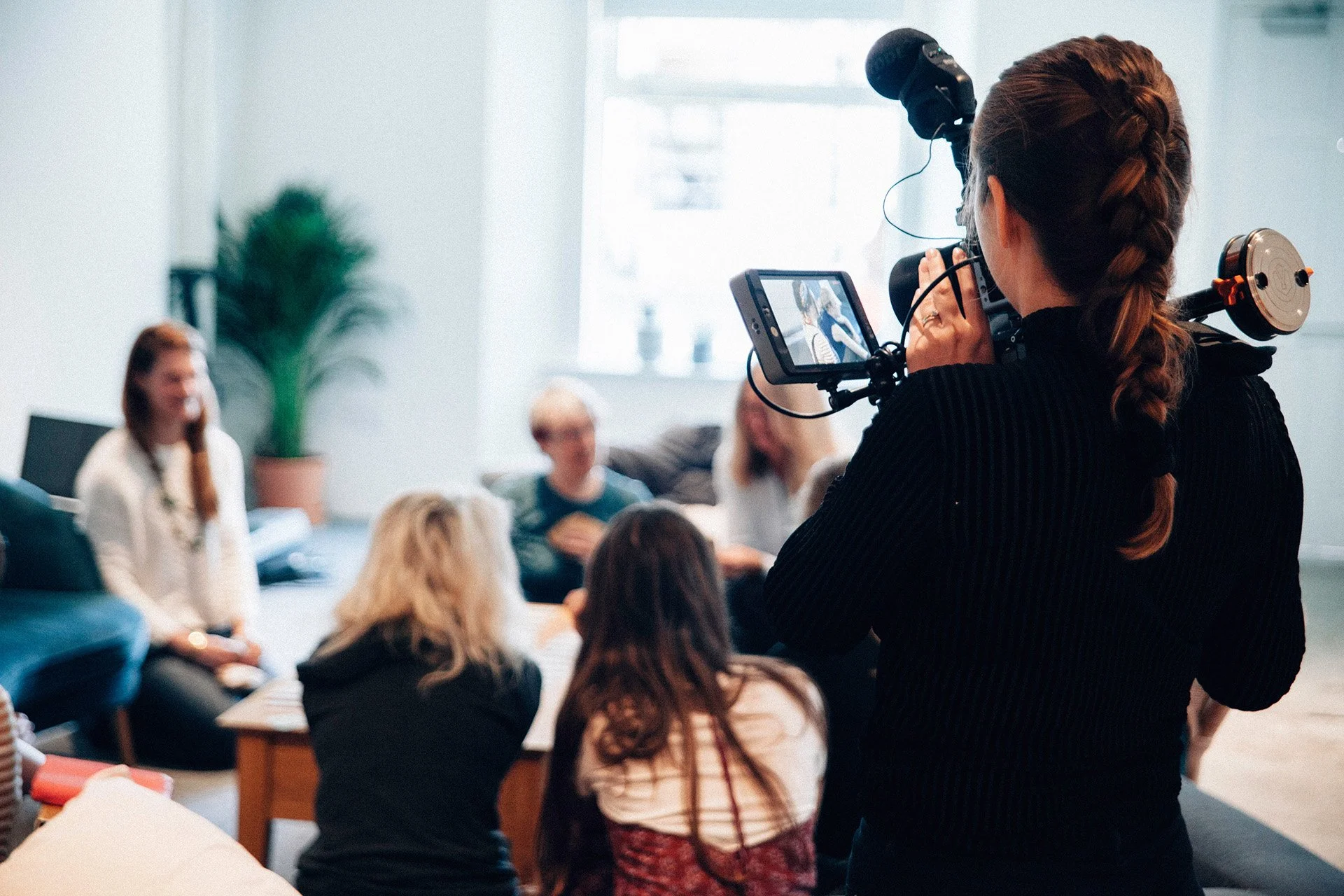 A woman with long, braided hair filming a group of people in a room with large windows and a potted plant.