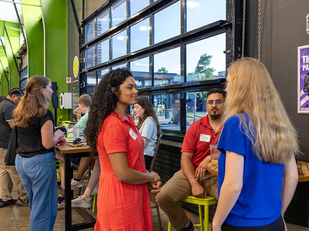 Two women are engaged in conversation in a lively indoor setting, with several other people at a counter in the background. Large windows reveal an outdoor scene with trees and buildings.