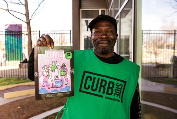 Man holding a magazine titled 'CURB' outside near a building, wearing a green vest with 'CURBSIDE' printed on it.