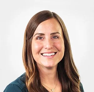Close-up portrait of a woman with long brown hair and a bright smile, wearing a dark teal top, against a white background.