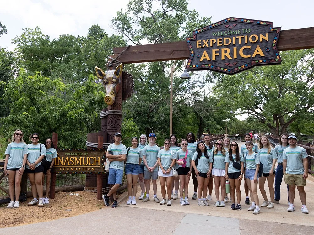 Group of people standing under a sign that reads 'Welcome to Expedition Africa,' with a decorative giraffe head sculpture and lush greenery in the background.