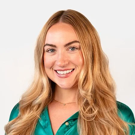 A young woman with long blonde hair smiling at the camera, wearing a teal satin shirt, against a plain light background.