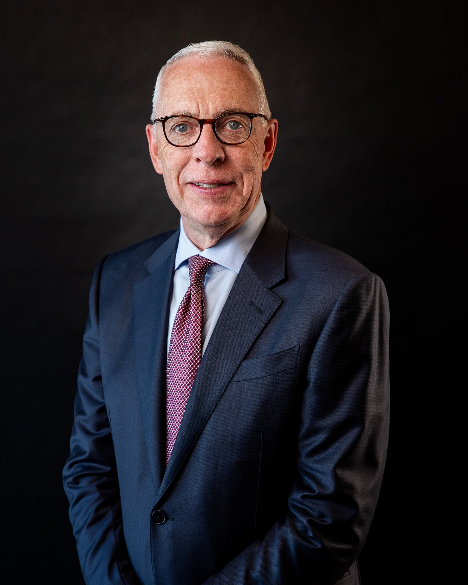 A smiling middle-aged man with short gray hair, glasses, wearing a blue suit jacket and light blue dress shirt, standing indoors with a projector screen in the background displaying the text 'GRIFFIN' and 'MEDIA'.