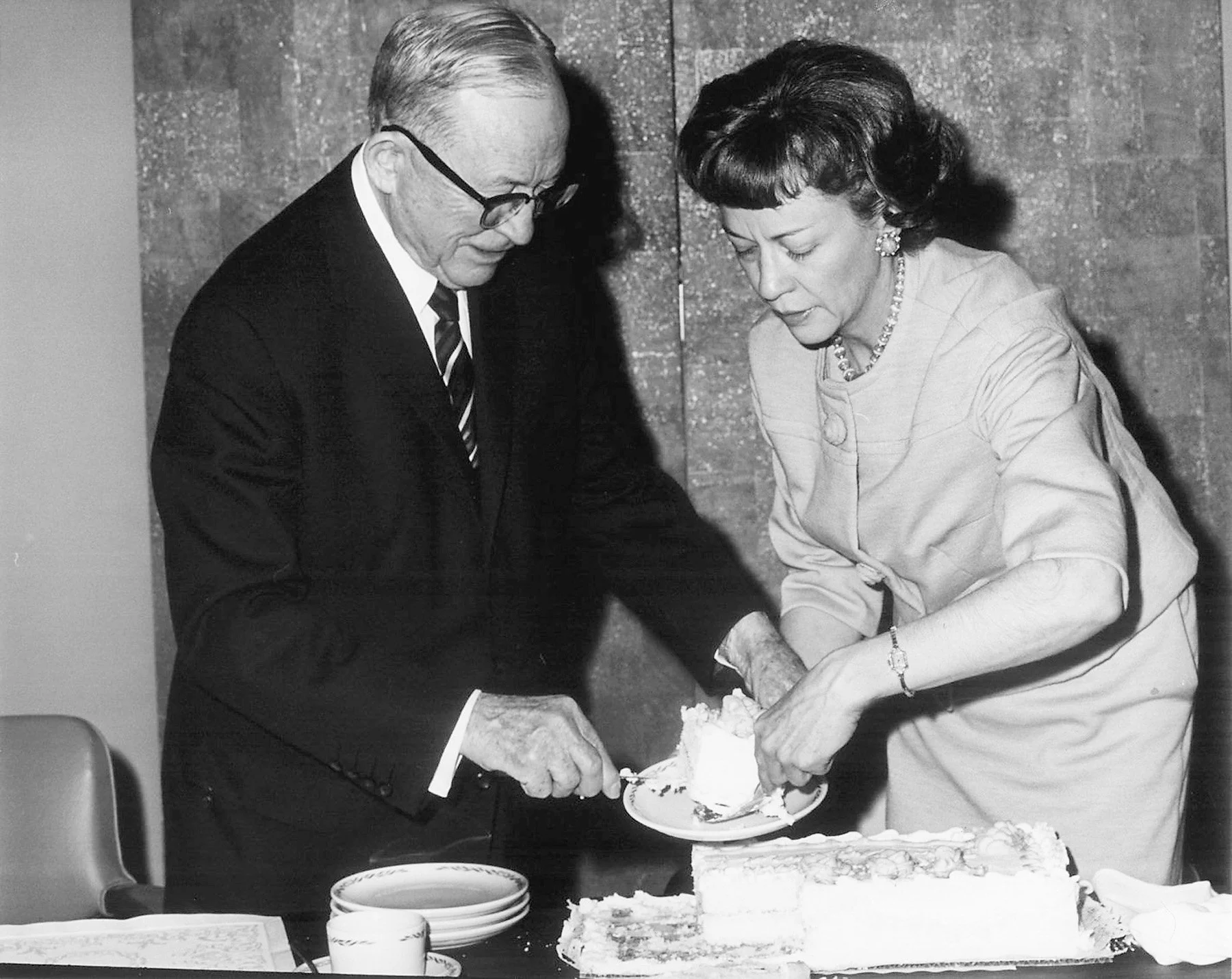 A black and white photo of a man and a woman cutting a cake. The man is wearing glasses, a suit, and a striped tie. The woman is dressed in a light-colored top accessories with jewelry, and has short hair. The focus appears to be on their hands as they cut the cake together, and there are plates and utensils on the table.