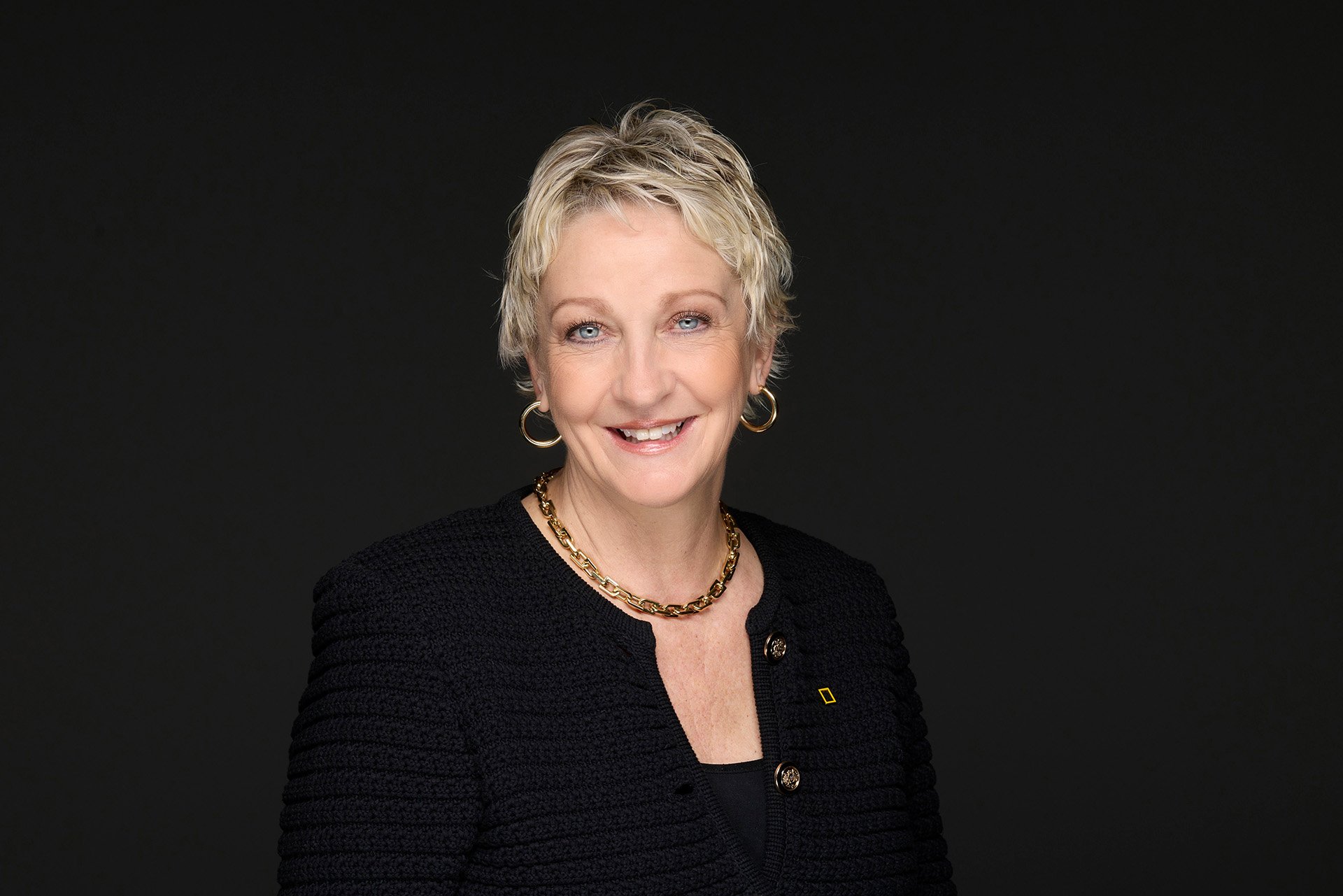 A professional woman with short blonde hair, wearing a black blazer over a white blouse, and gold hoop earrings, against a dark background.