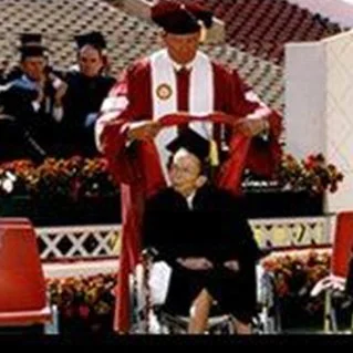 Individual in academic regalia reading a diploma to a seated graduate at a graduation ceremony.