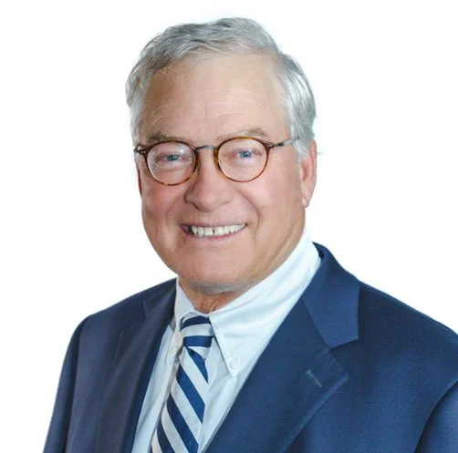 A smiling older man with glasses, gray hair, wearing a navy suit and a striped tie, against a white background.