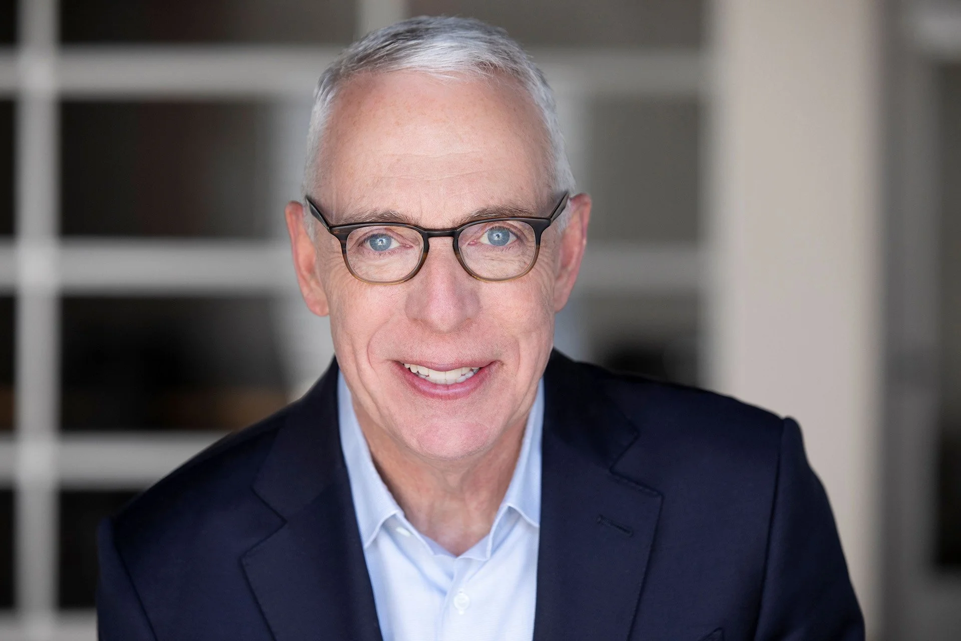 A smiling middle-aged man with short gray hair, glasses, wearing a blue suit jacket and light blue dress shirt, standing indoors with a projector screen in the background displaying the text 'GRIFFIN' and 'MEDIA'.