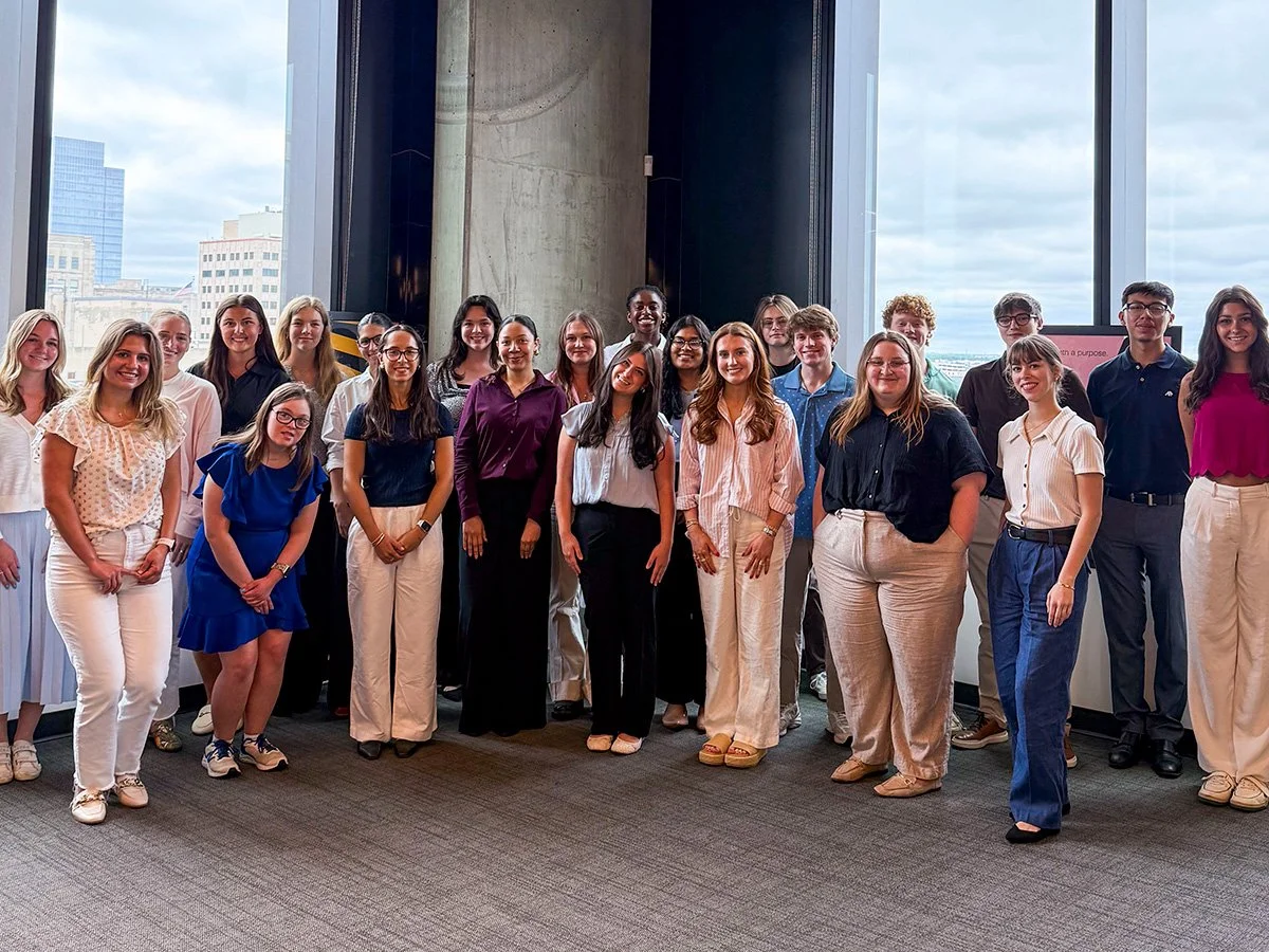 Group of diverse young adults, both men and women, standing indoors in front of large windows with city skyline view, dressed in casual and business casual attire.