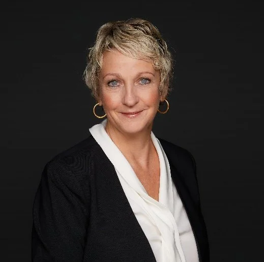 Portrait of a middle-aged woman with short blonde hair, blue eyes, wearing a black blazer over a white blouse, gold hoop earrings, and smiling against a dark background.