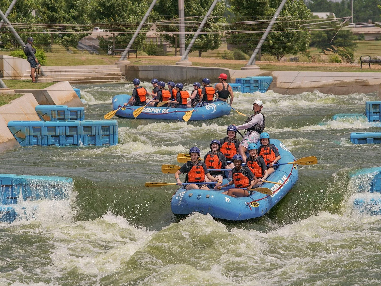 People white water rafting in an outdoor river, wearing life jackets and helmets, with some smiling and paddling.
