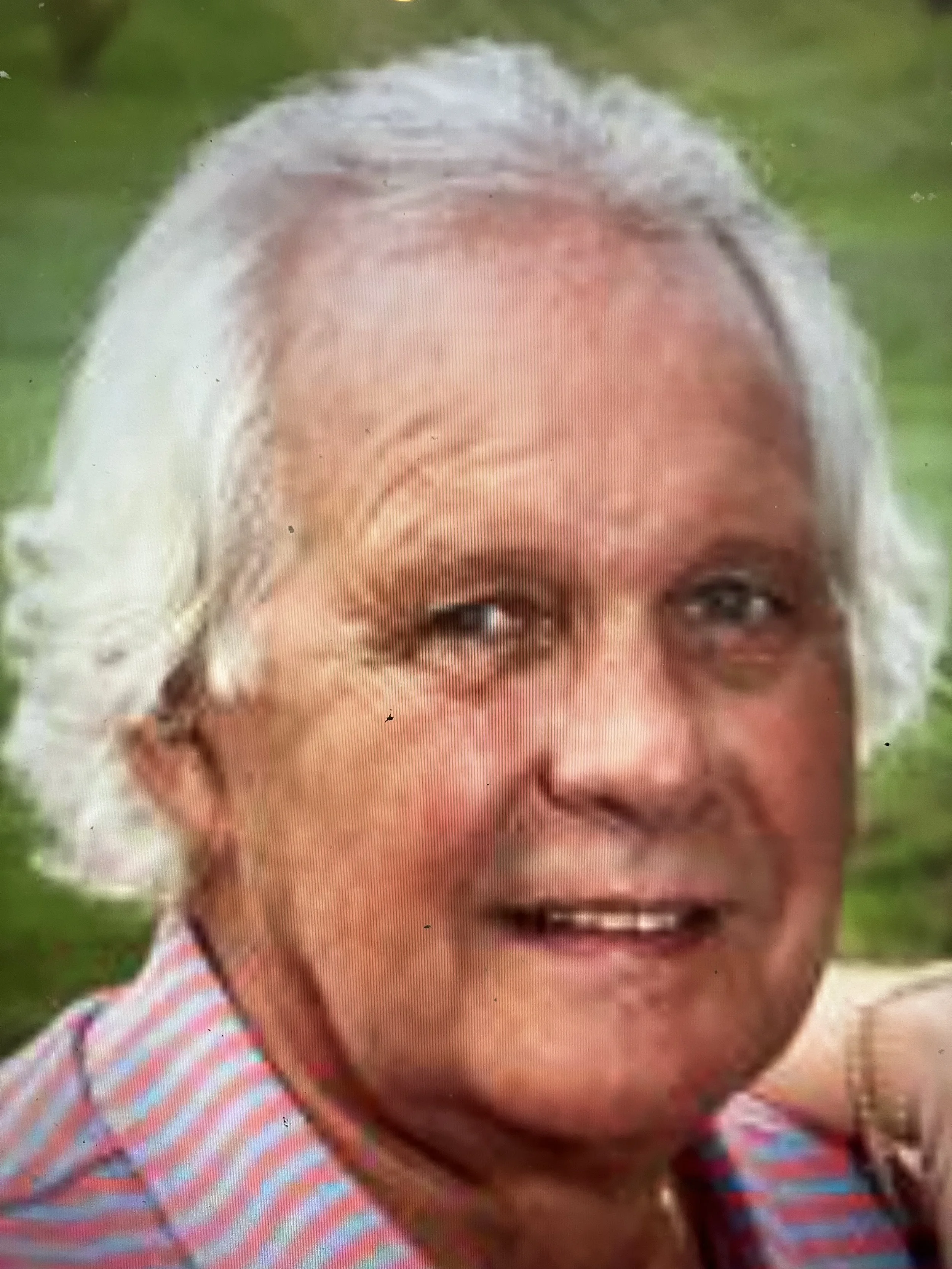 Close-up of an elderly woman with gray hair, wearing a striped shirt, outdoors with greenery in the background.