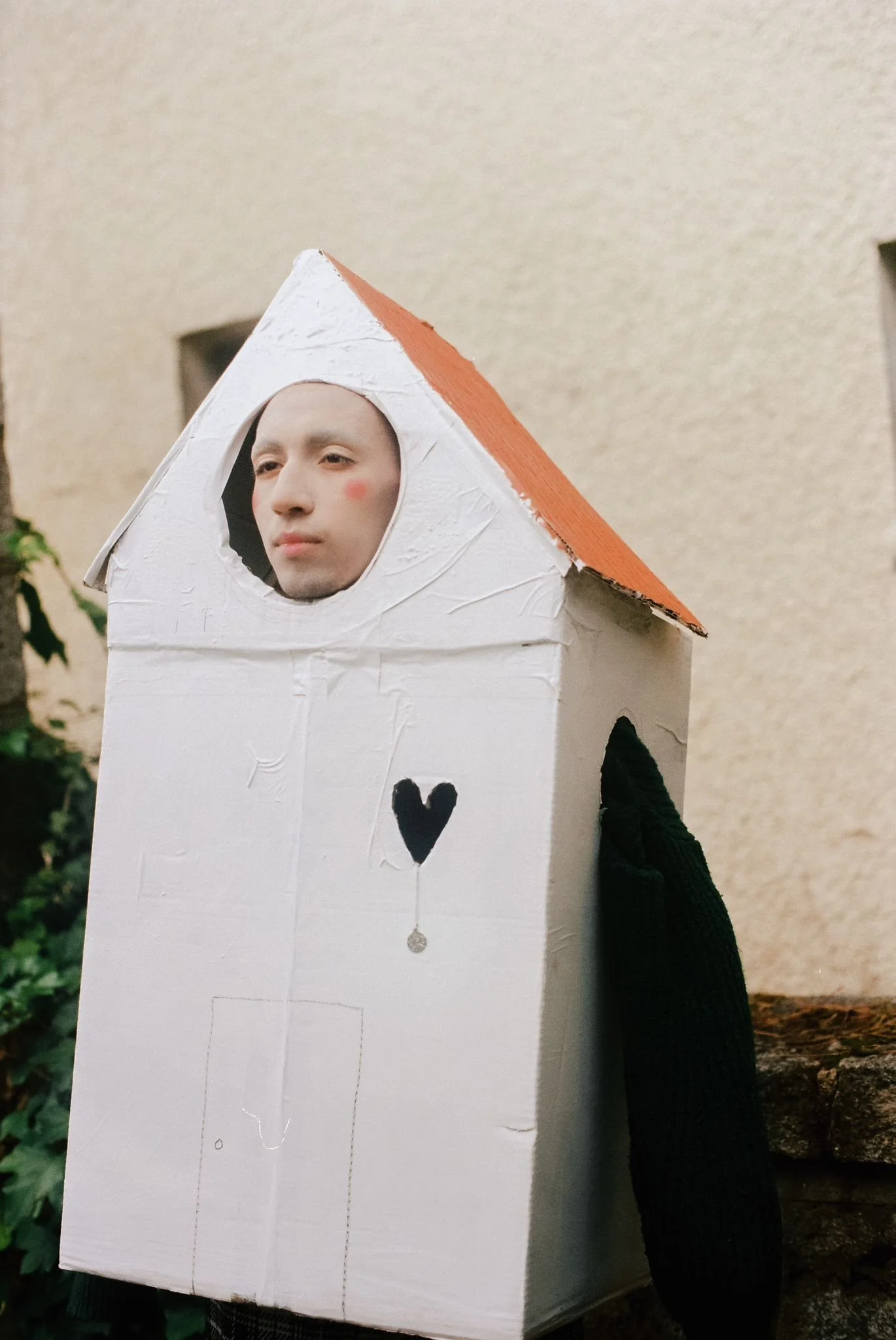 Persona con rostro de expresión neutral dentro de una casa de cartón pintada de blanco y con techo de tejas, con un corazón negro y una gota de agua en la fachada