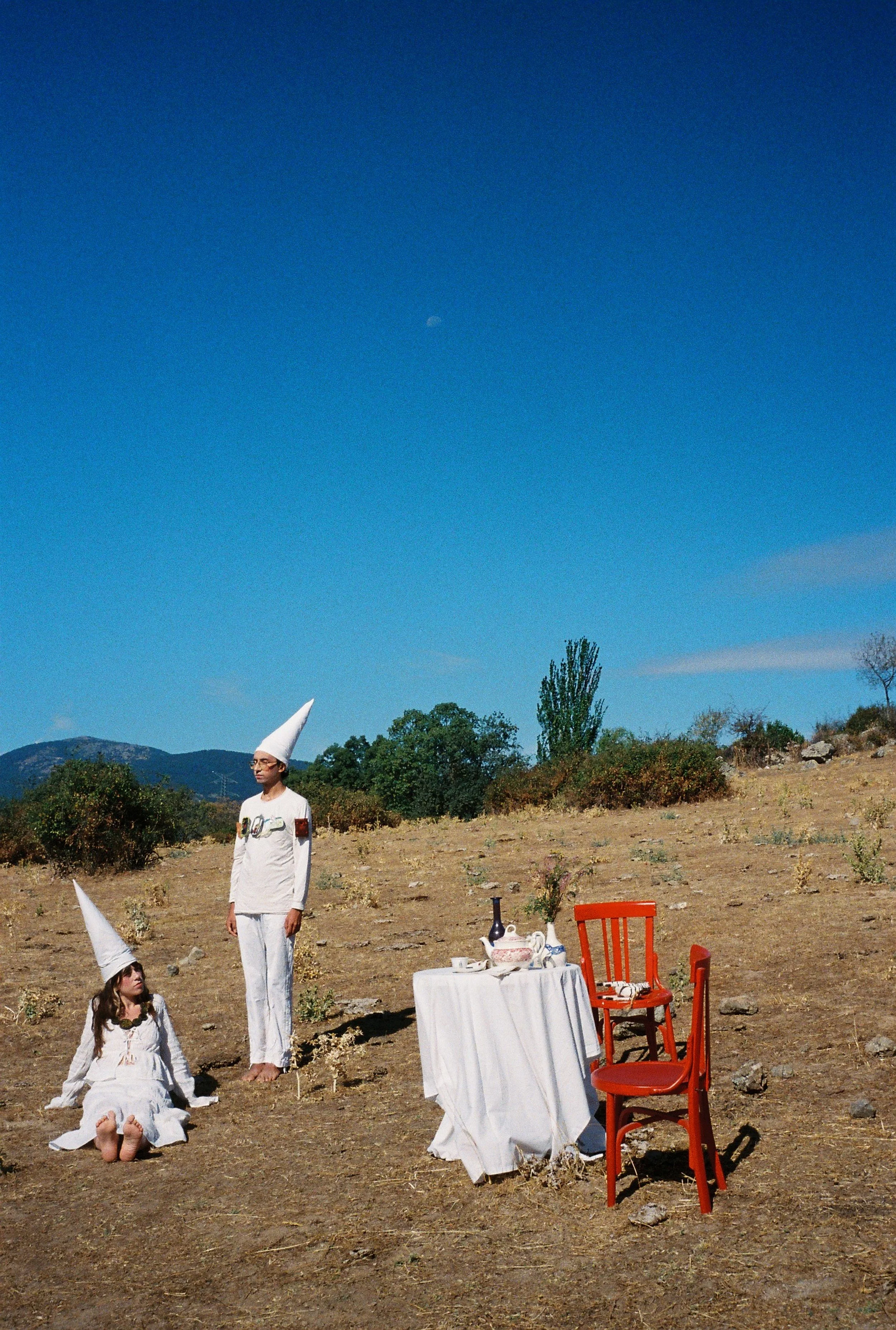 Una escena surrealista en un campo abierto con cielo azul y luna visible, con dos figuras humanas en trajes blancos con gorros con forma cónica, una de pie y otra sentada en el suelo. Una mesa cubierta con un mantel blanco, con vajilla y una botella, y dos sillas rojas frente a ella.