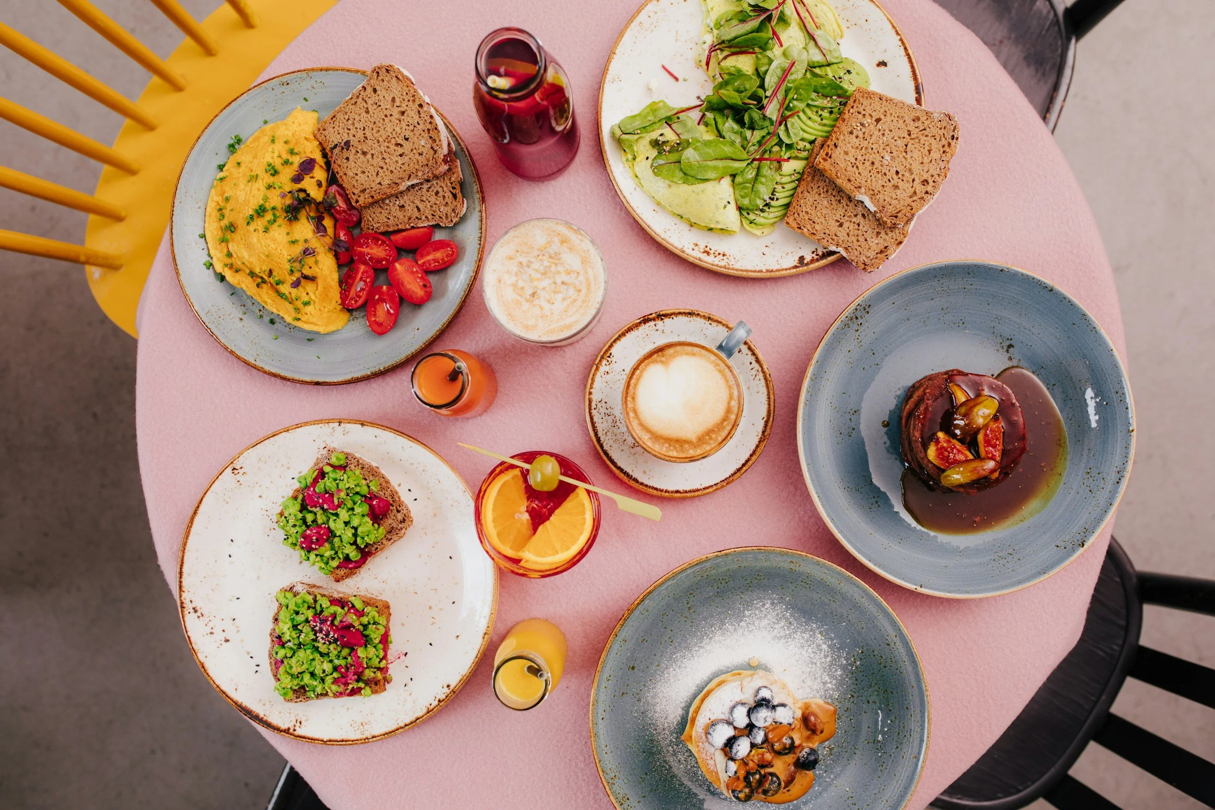 A top-down view of a pink table with various breakfast dishes, including scrambled eggs with cherry tomatoes and toast, a green salad, two pieces of bread, a dessert with blueberries and caramel, a glass of orange juice with a cherry, a coffee mug with cappuccino, a small cup of whipped cream, small bottles of juice, and a cooked dish in a blue bowl.