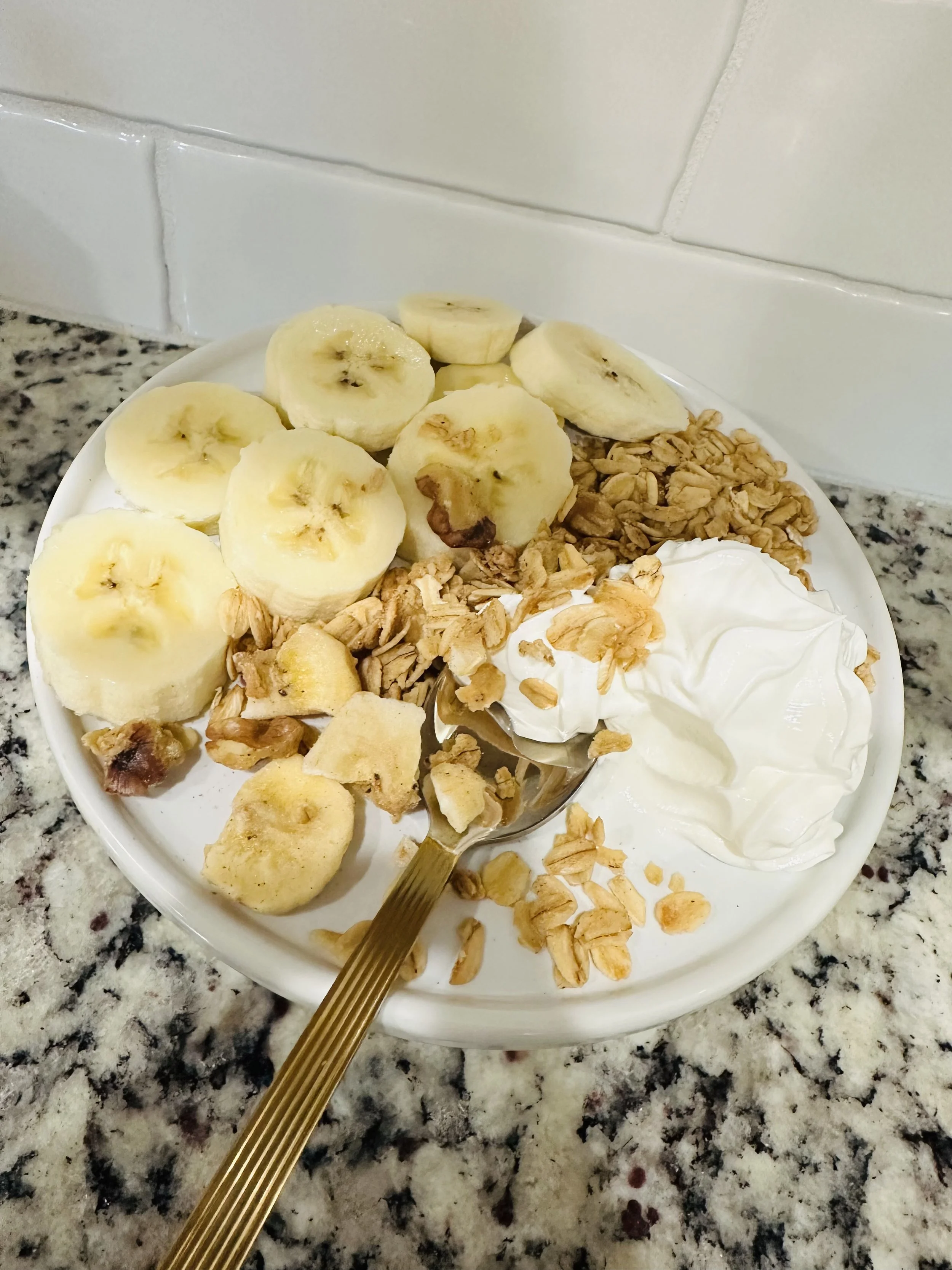 Banana slices, granola, yogurt, and granola on a white plate on a granite countertop.