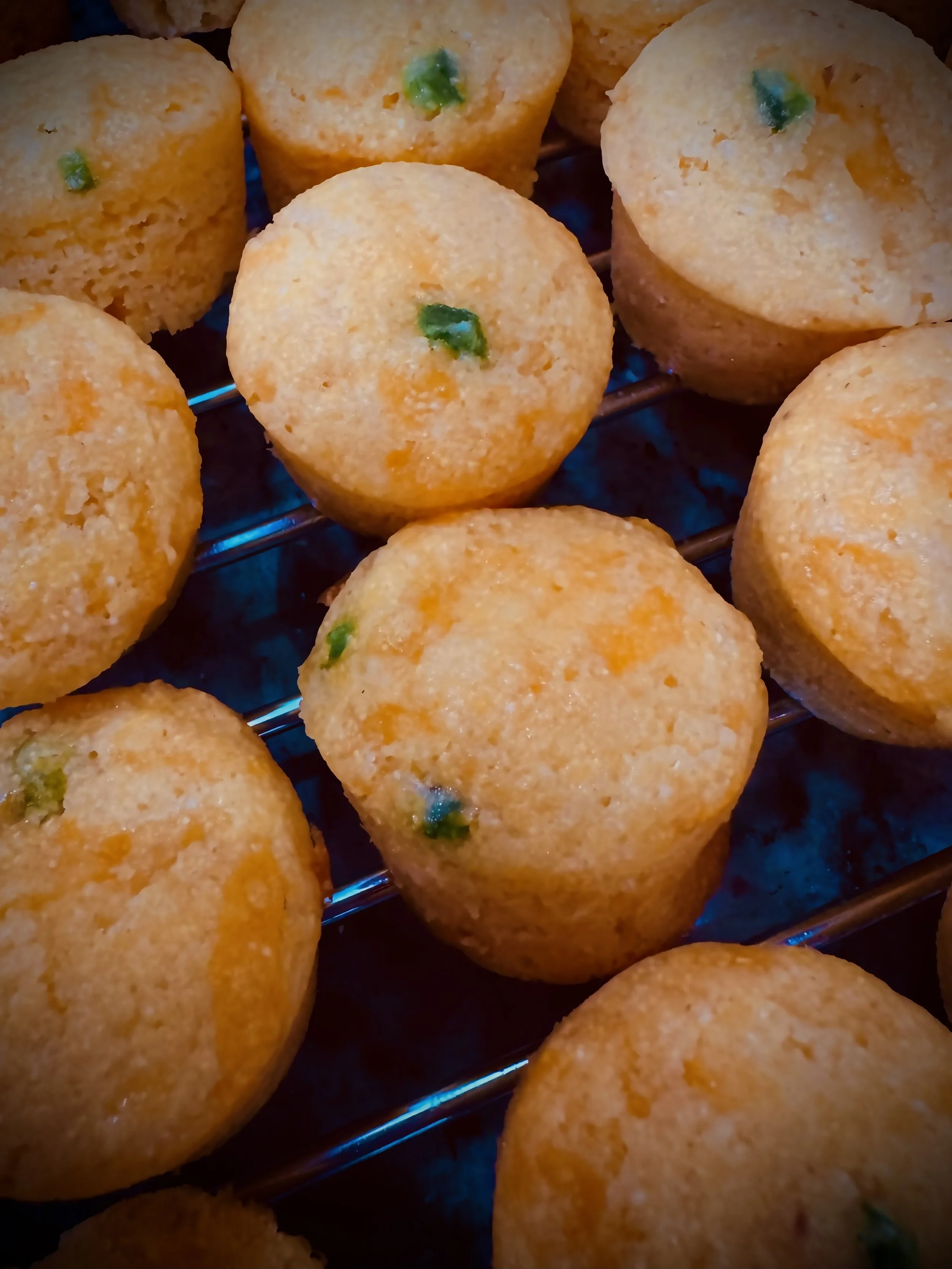A close-up view of cornbread muffins on a wire cooling rack, garnished with small green chili pieces.