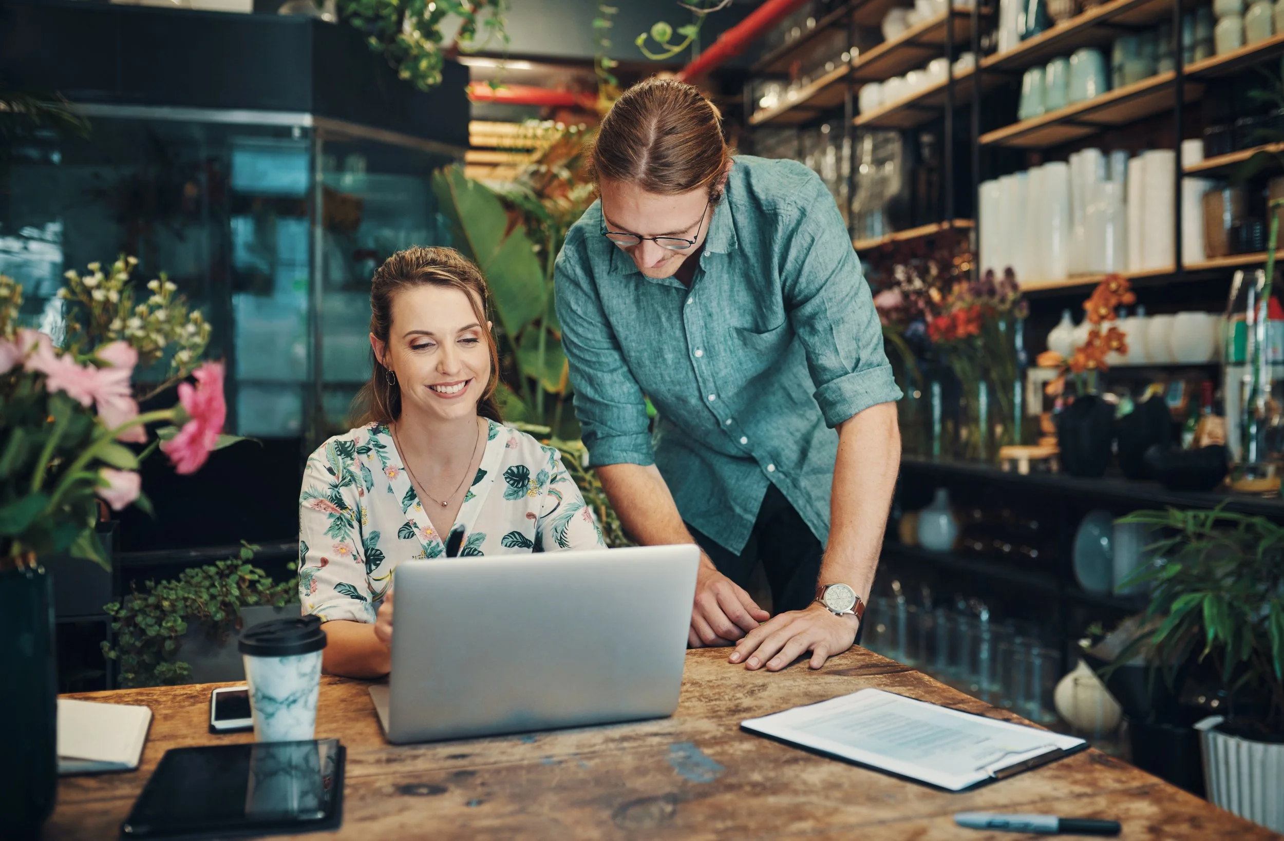 Una mujer sonriente con cabello rizado sentado frente a una laptop en una mesa de madera en un ambiente de cafetería o librería, con un hombre de cabello largo y gafas de pie al lado, ambos revisando la pantalla.