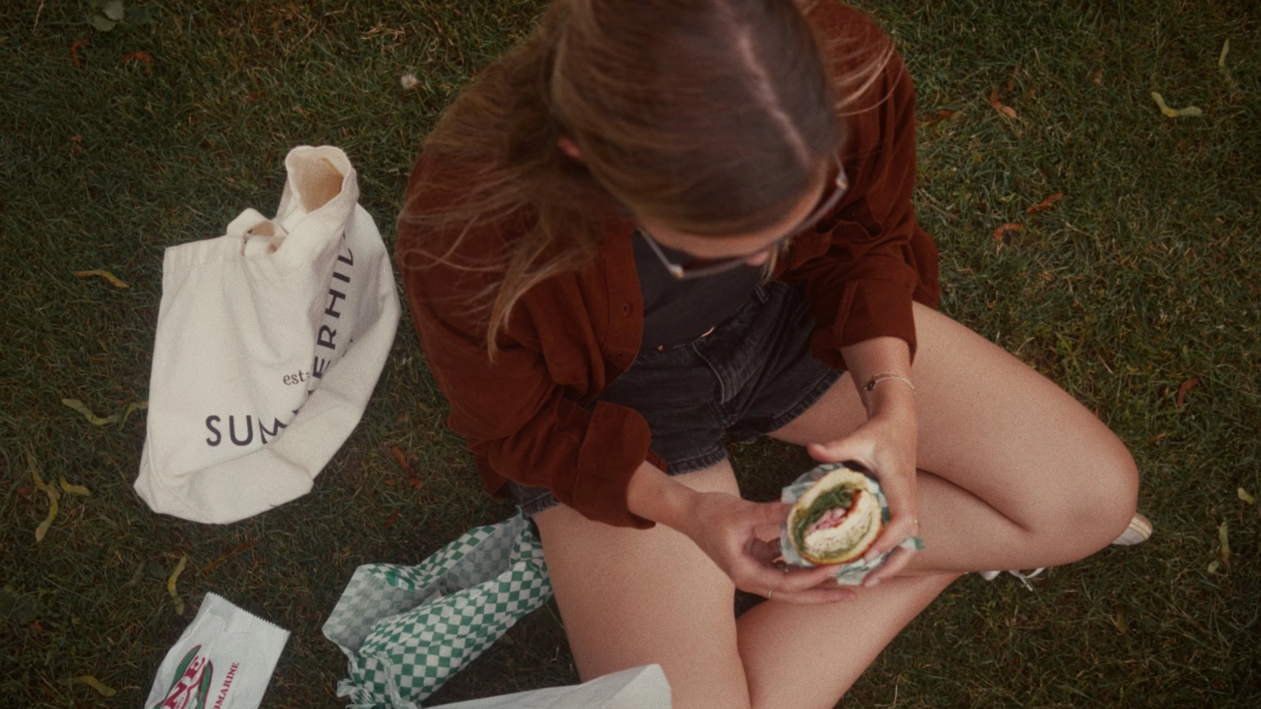 A person sitting on the grass holding a sandwich, with a white bag and other items nearby.