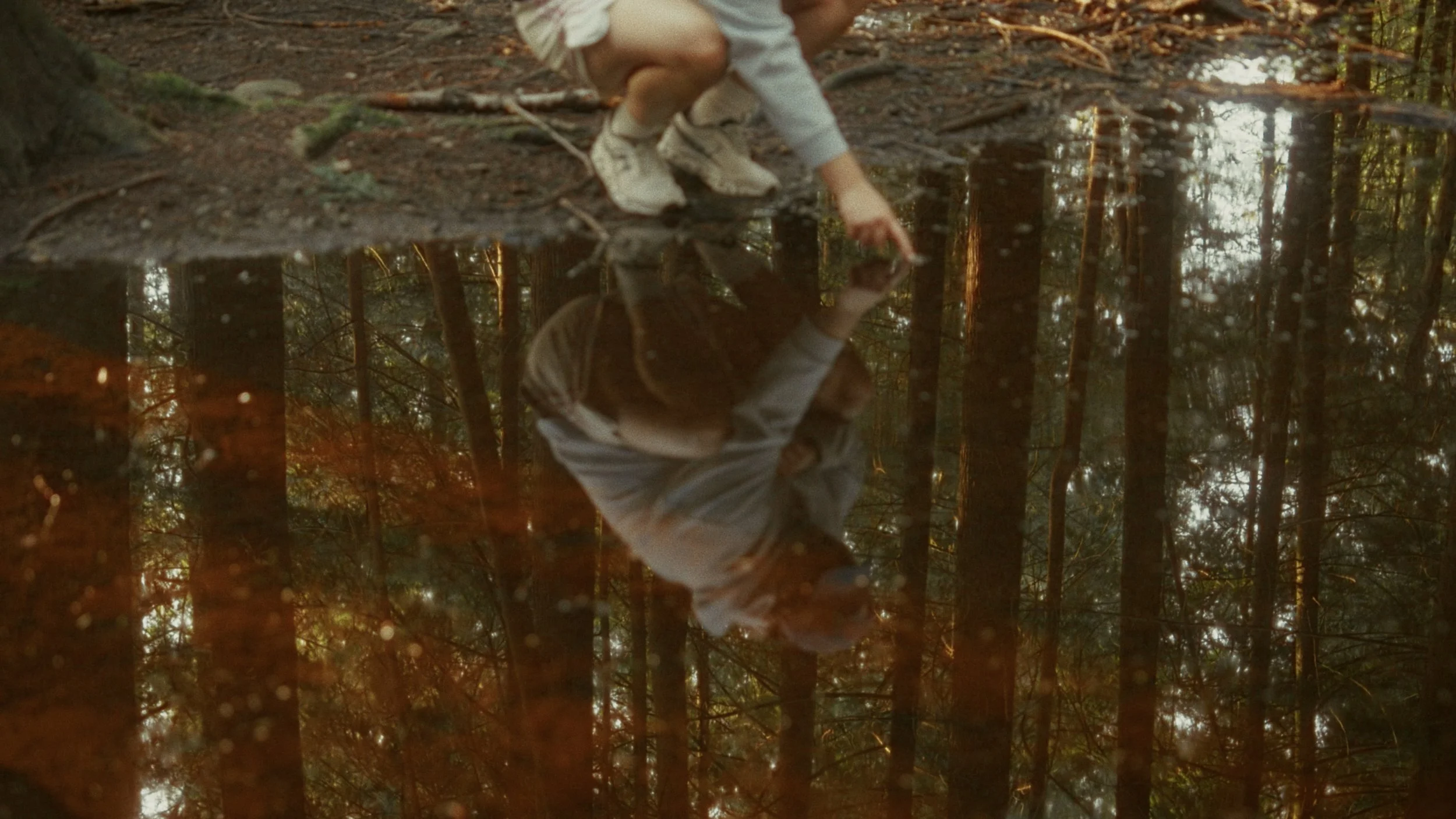A young person crouching by a small pond in a forest, touching the water's surface, with trees and foliage reflected in the water.
