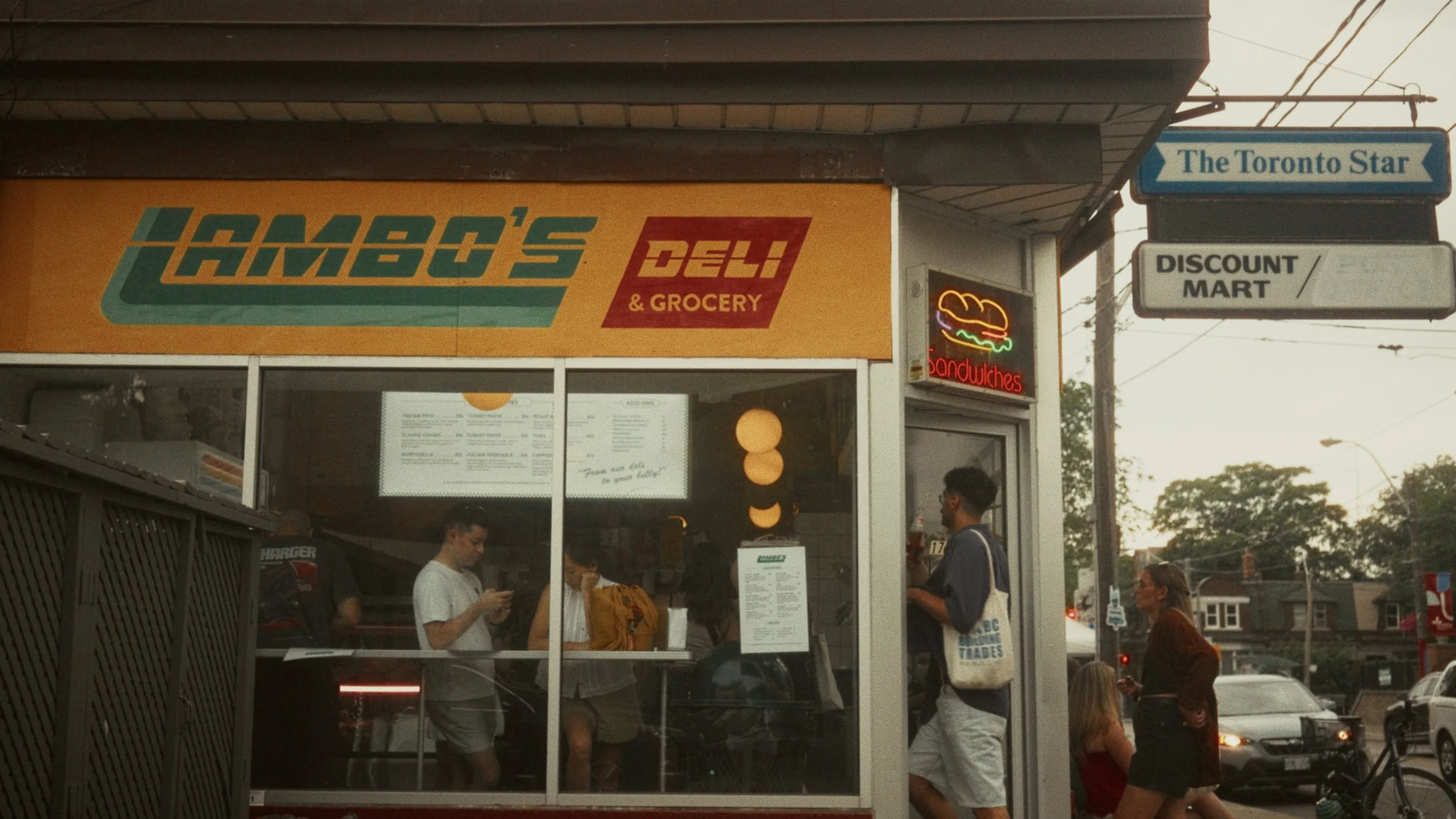 Exterior view of James' Deli & Grocery with customers inside and outside, neon sandwich sign, and street scene in Toronto.