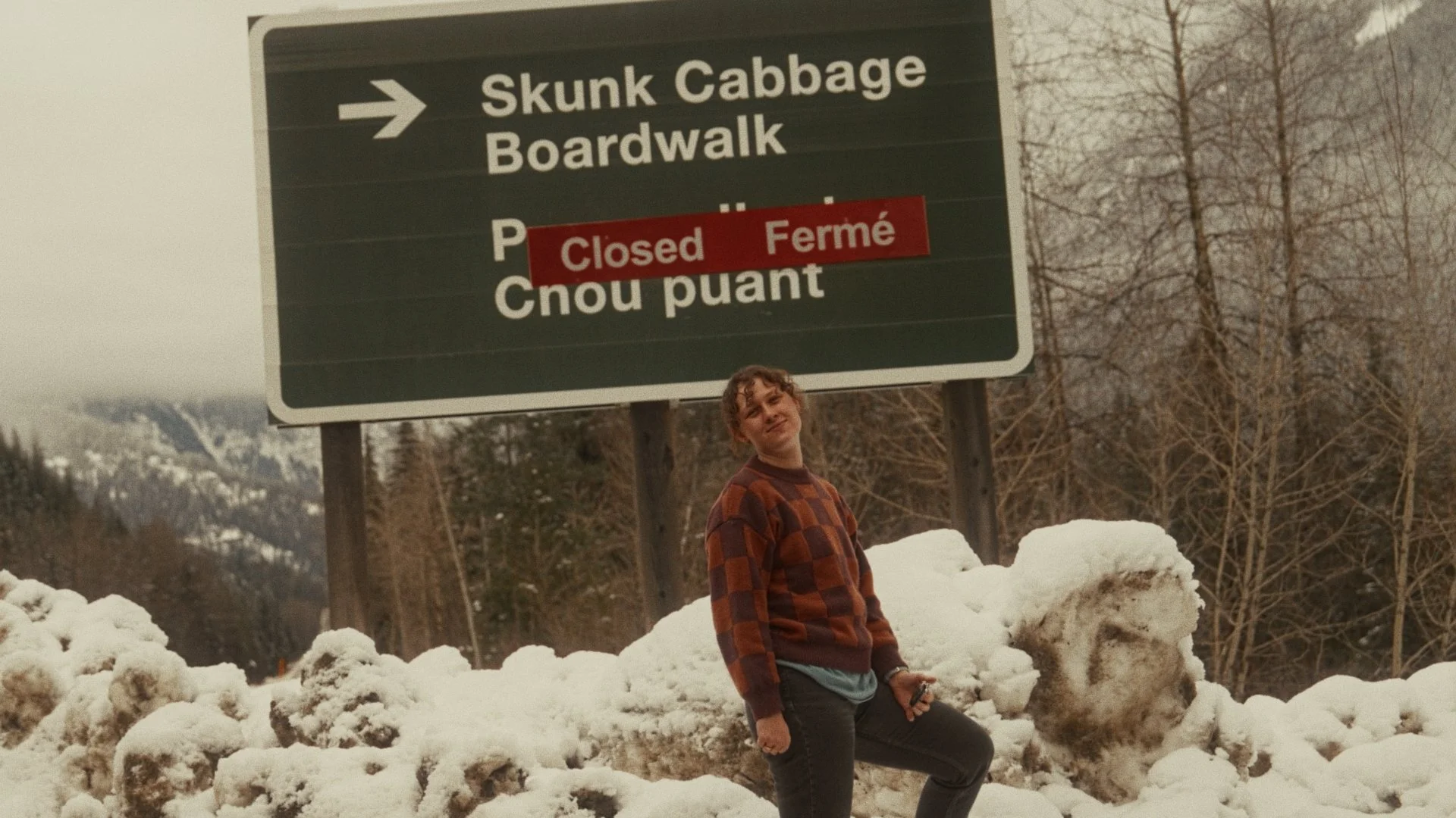 A young woman standing in front of a large green road sign with a red "Closed" sticker. The sign indicates directions to Skunk Cabbage Boardwalk and other locations. She is outdoors in a snowy area with trees and mountains in the background, wearing a brown and orange checkered sweater and dark pants.