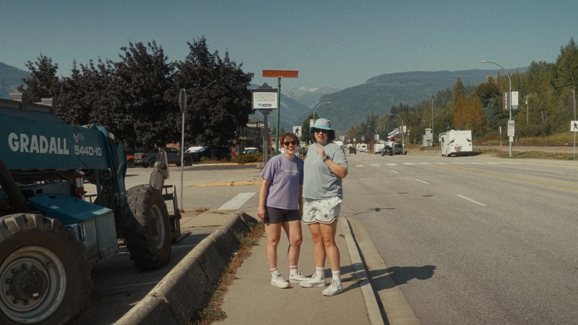 Two women standing on a sidewalk near a street with cars traveling in the background, mountains, and trees behind them. One woman is wearing a purple shirt, black shorts, white socks, and sneakers, while the other is wearing a gray shirt, shorts, a hat, and sneakers.