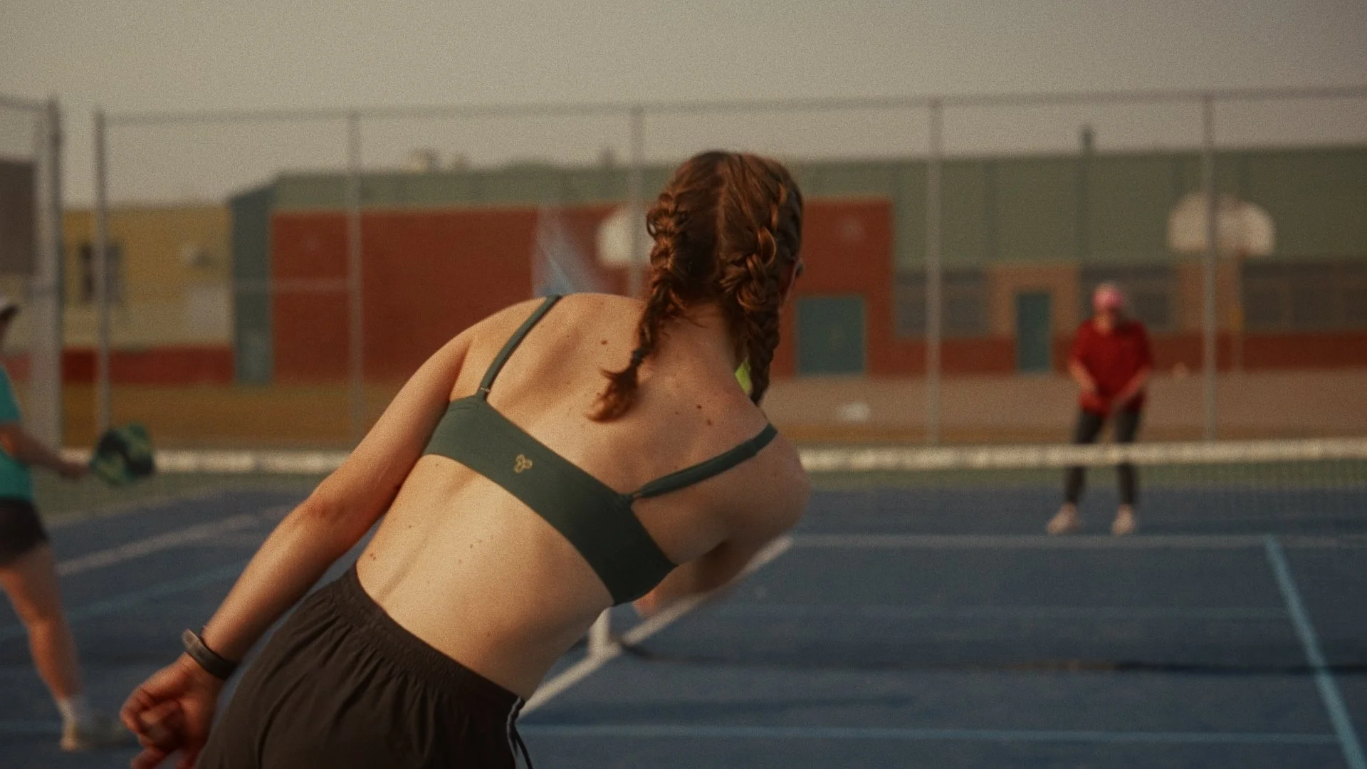 A girl with braided hair in athletic clothing serving a volleyball during a game on an outdoor tennis court.