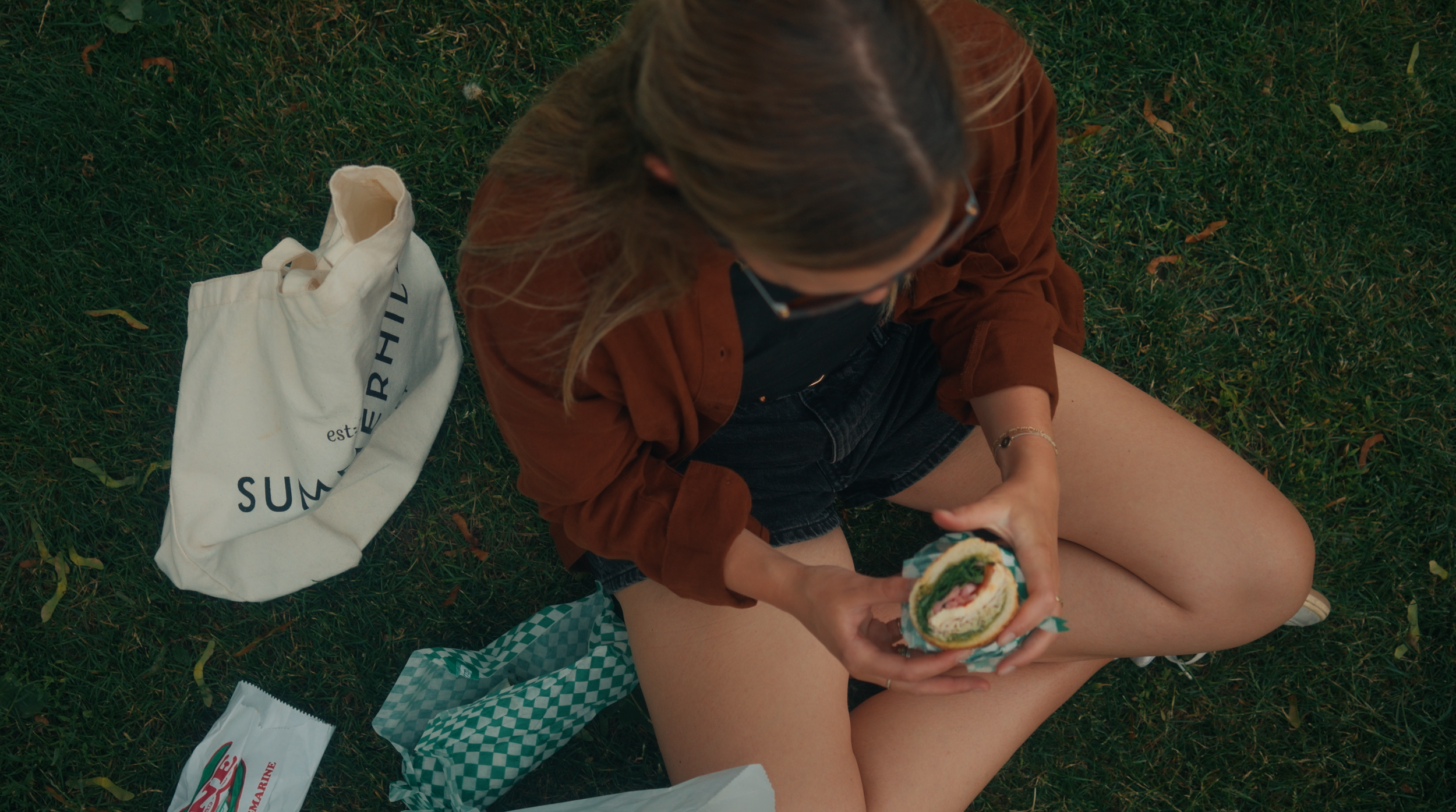 A young woman sitting on grass with picnic items around her, holding a sandwich with lettuce, tomato, and cheese inside.