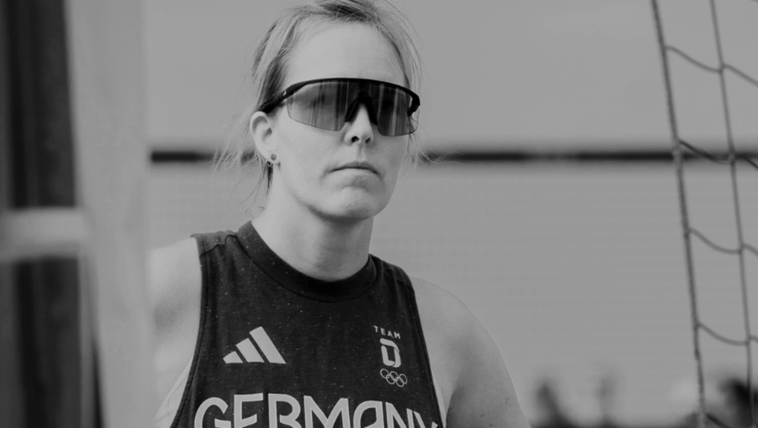 Schwarz-Weiß-Foto von Katharina Hubert, Physiohub Hamburg, die eine dunkle Sportbrille trägt, im Trikot mit Deutschland-Flagge und Olympische Ringe, vor einem Volleyballnetz.