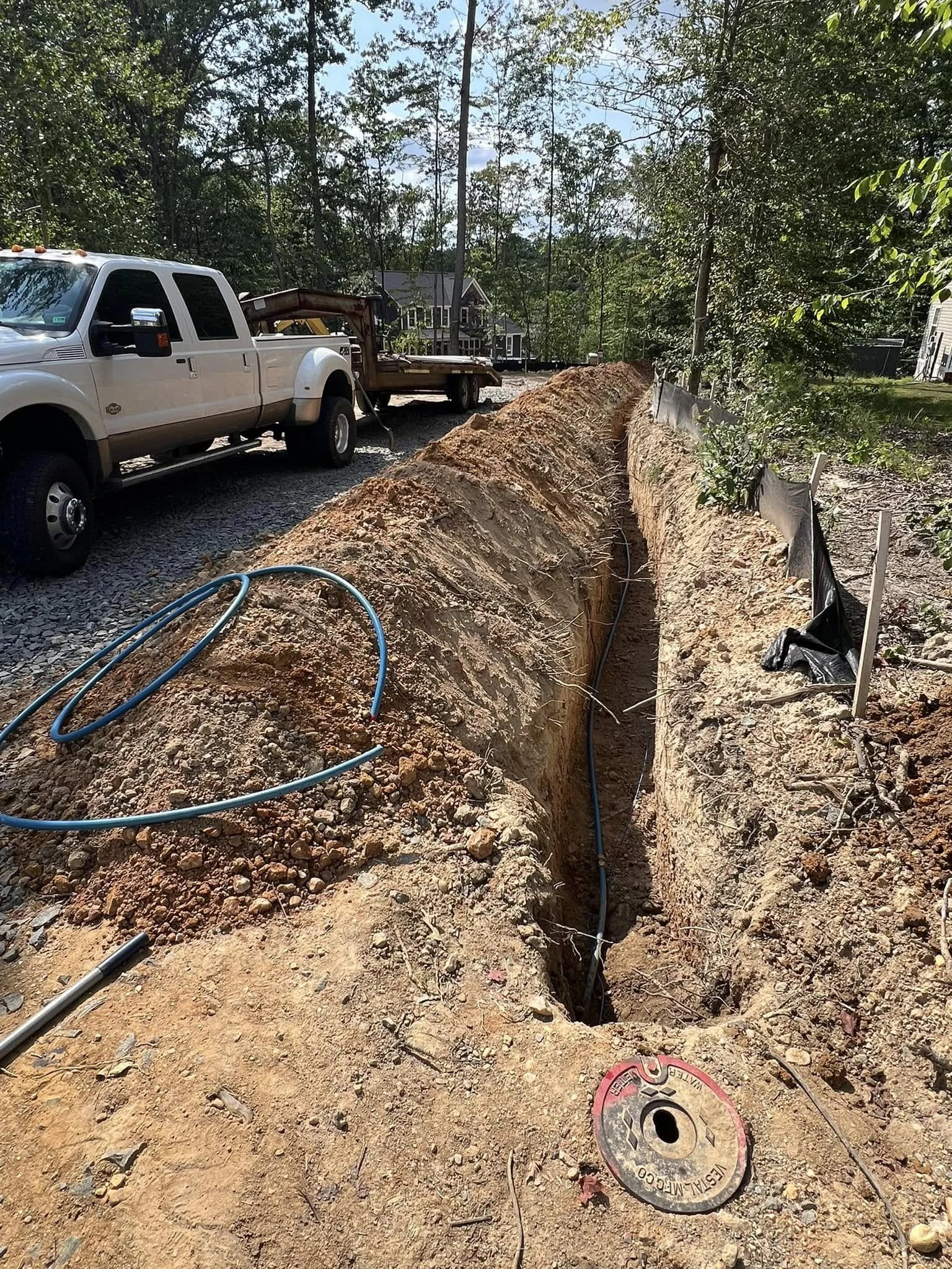 Construction site with a deep trench dug into the ground, featuring electrical or plumbing cables inside, surrounded by dirt and gravel, with a white pickup truck and a flatbed trailer nearby, and trees and houses in the background.